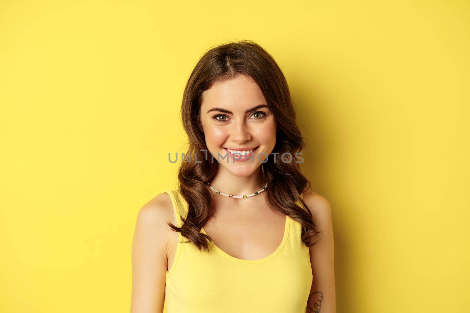 Close up portrait of brunette woman with healthy white smile, looking happy and confident, posing against yellow background.