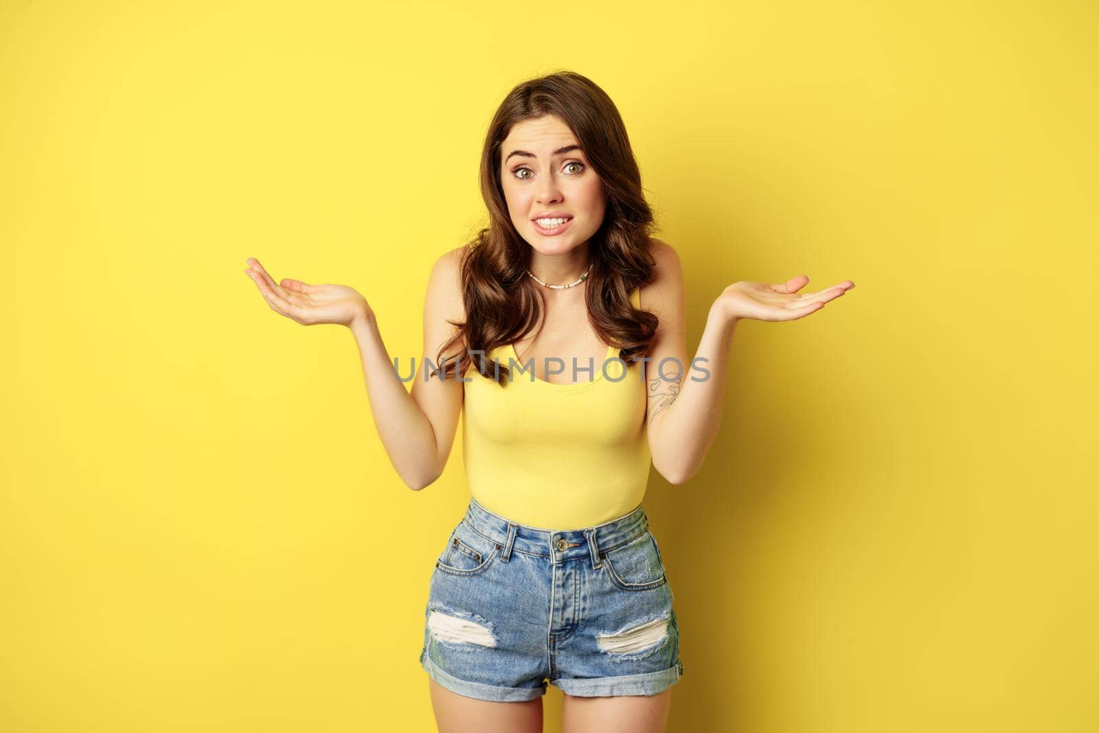 Woman shrugging shoulders and looking clueless, apologizing and smiling awkward, dont know, puzzled, has no idea, standing against yellow background by Benzoix