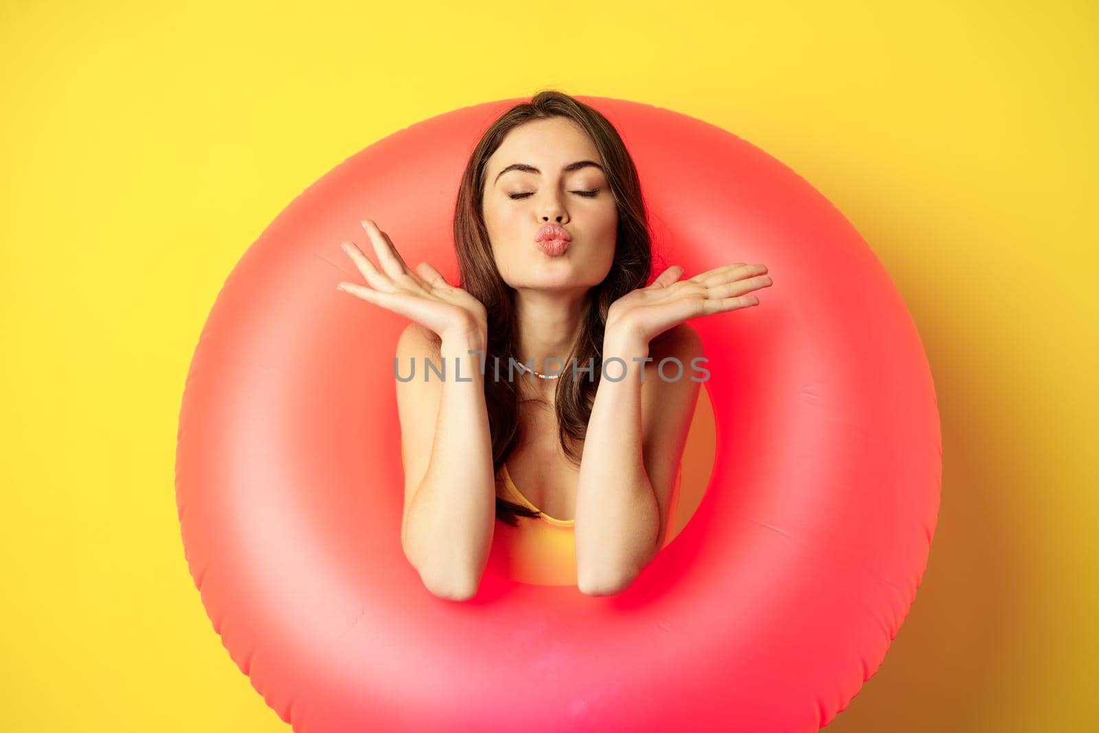 Beautiful coquettish woman inside pink swimming ring, posing on summer vacation, beach holiday concept, standing against yellow background.