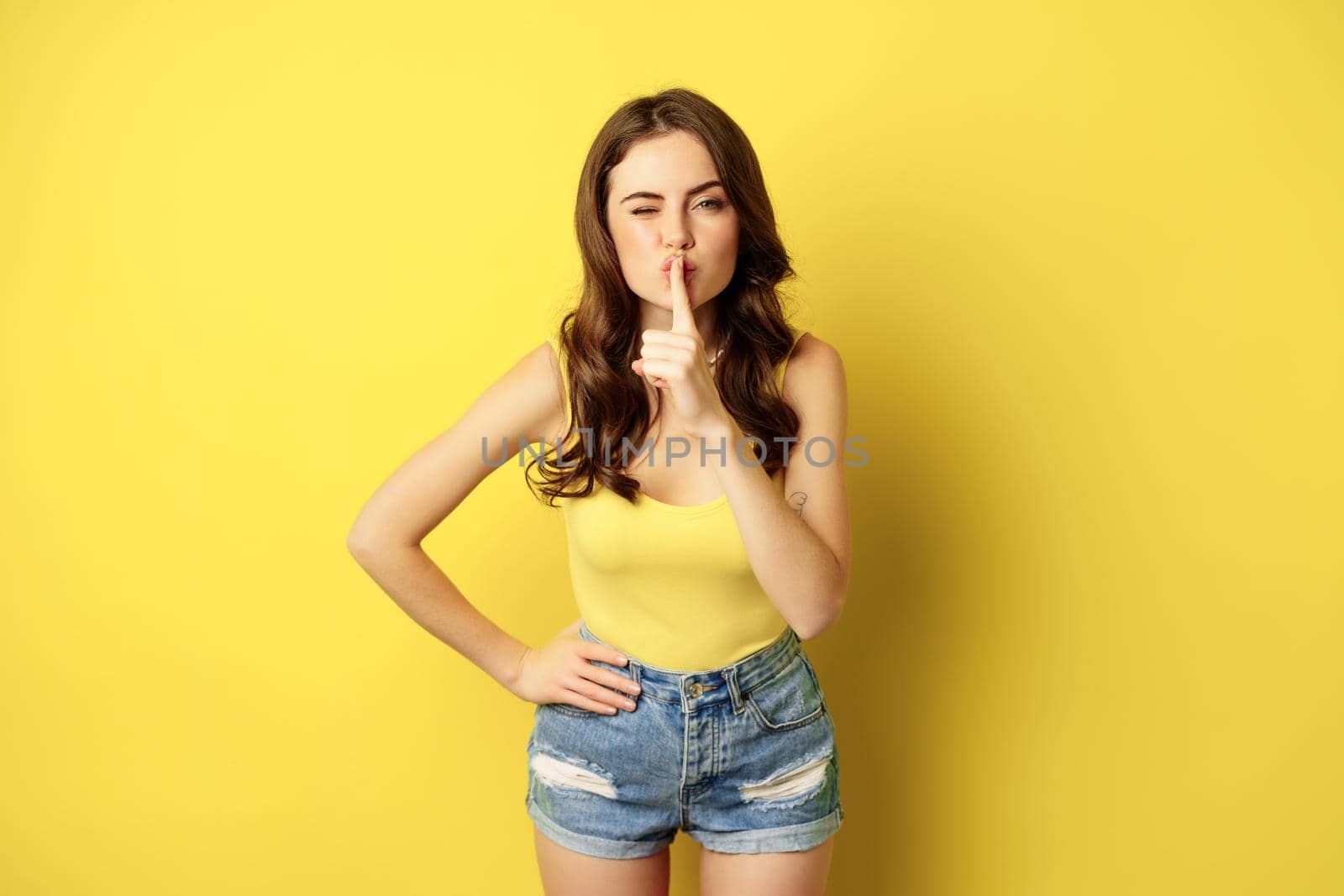 Young woman keeps secret, shows silence quiet, shush gesture, hushing, press finger to lips, standing over yellow background. Copy space