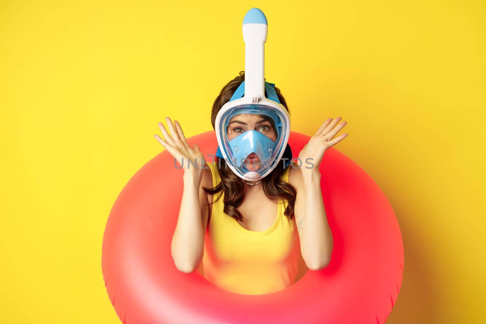 Portrait of enthusiastic young woman on vacation, wearing snorkling, diving mask and swimming ring, standing over yellow background.