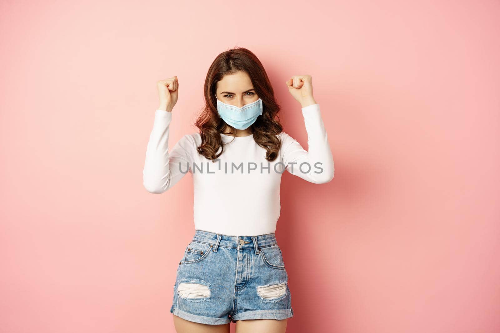 Enthusiastic brunette girl in medical face mask, dancing and laughing, celebrating victory, triumphing, winning smth, standing over pink background.