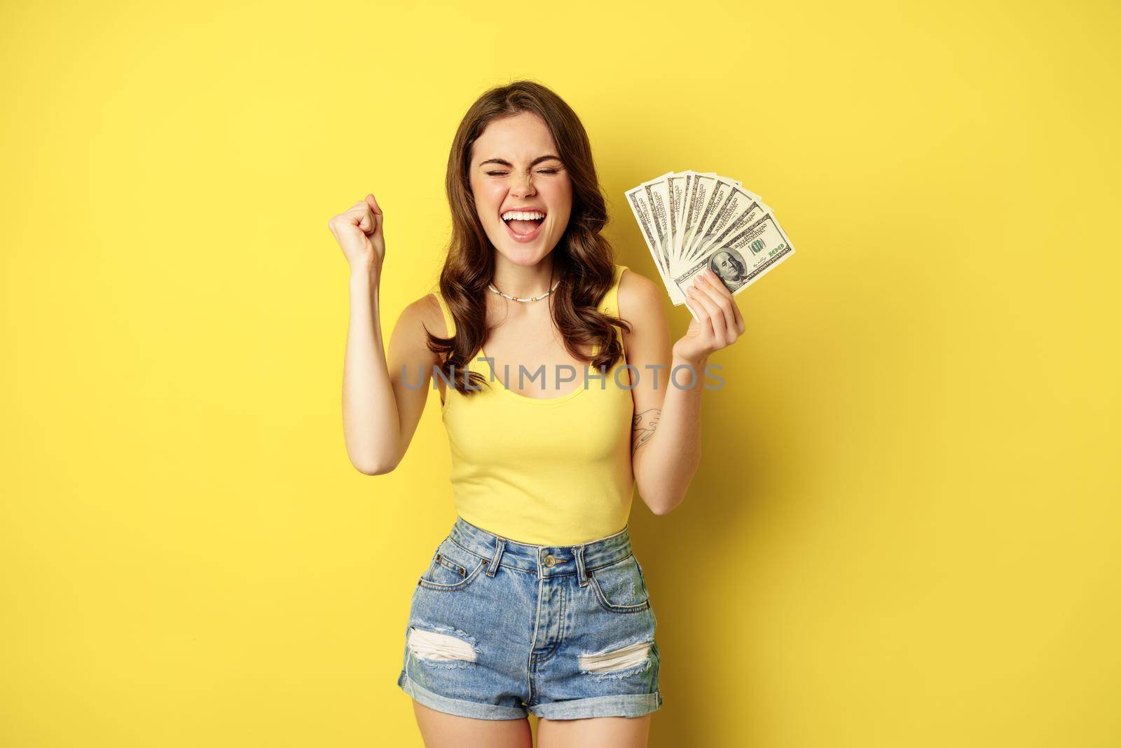 Enthusiastic smiling woman showing money cash, laughing and looking excited, standing in summer clothes against yellow background.