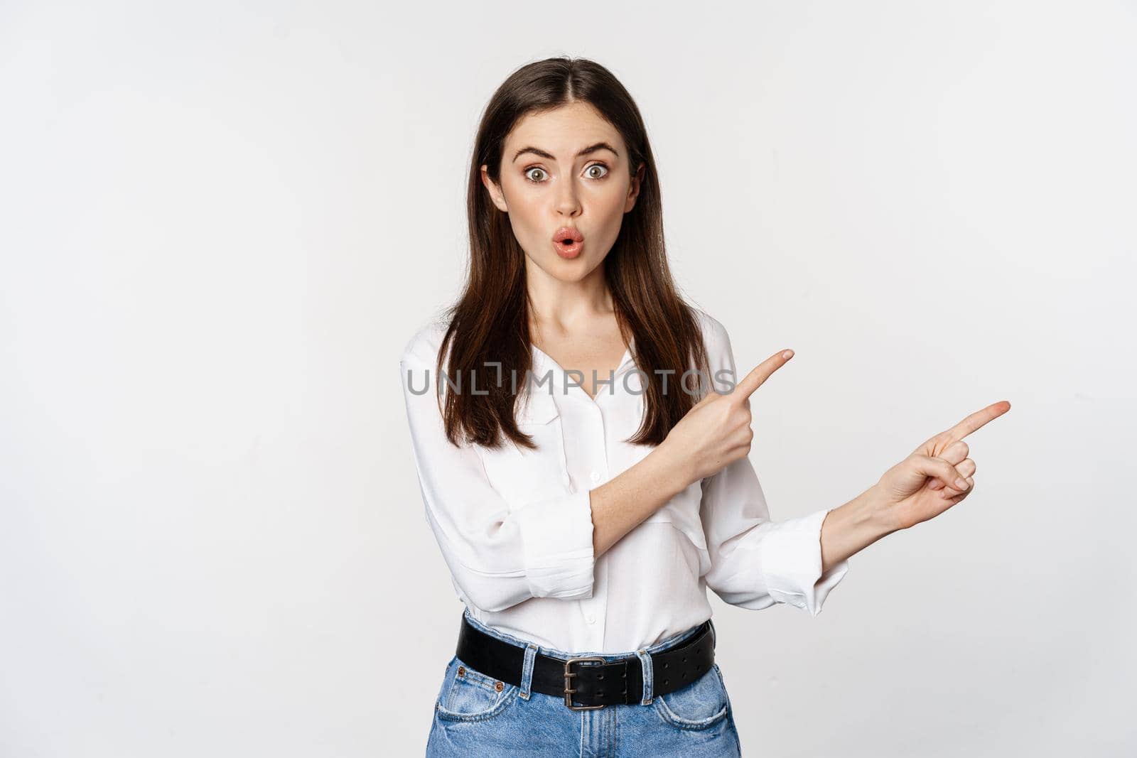 Amazed young woman pointing fingers down, showing announcement or logo banner, looking surprised and intrigued, standing over white background.