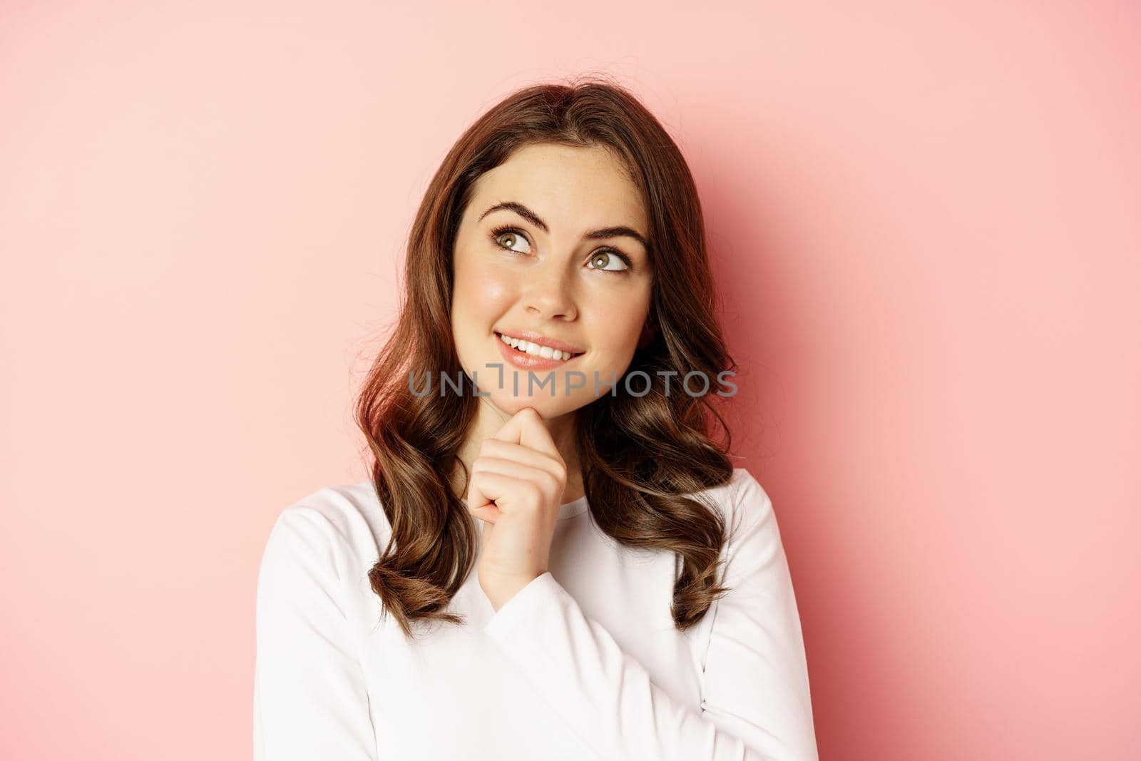 Close up portrait of coquettish smiling woman, glamour girl thinking, looking thoughtful, standing over pink background.