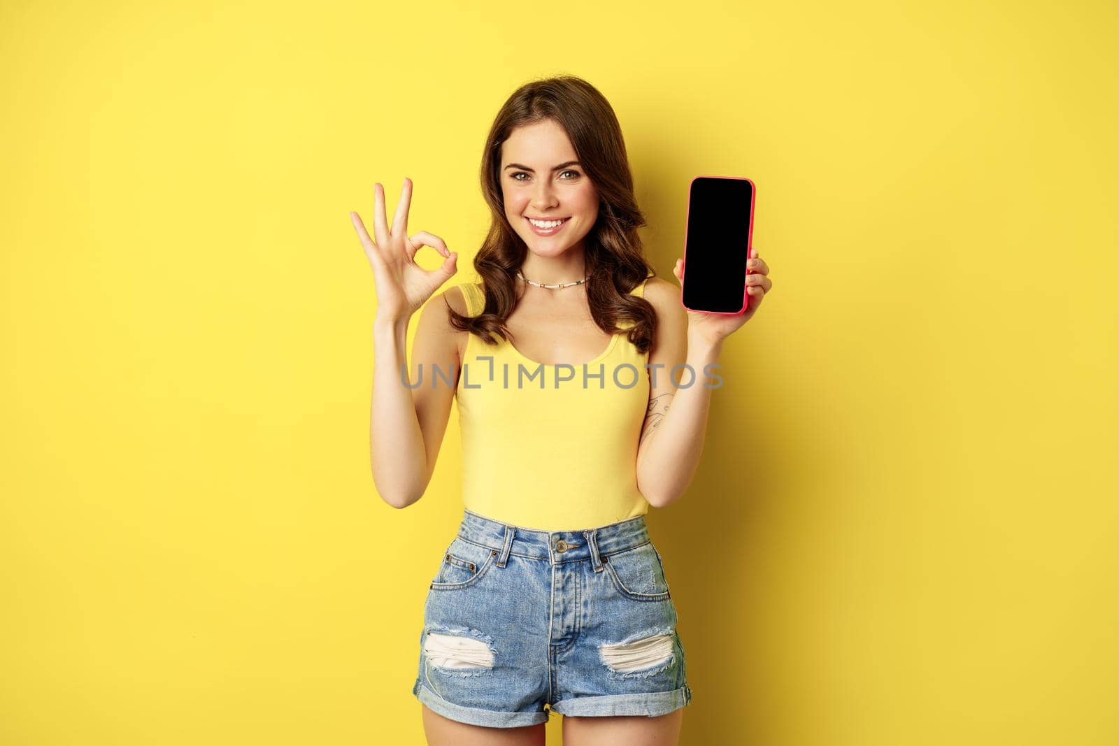 Pretty brunette woman, showing mobile phone screen, smartphone app interface and okay sign, recommend website, store, standing against yellow background.