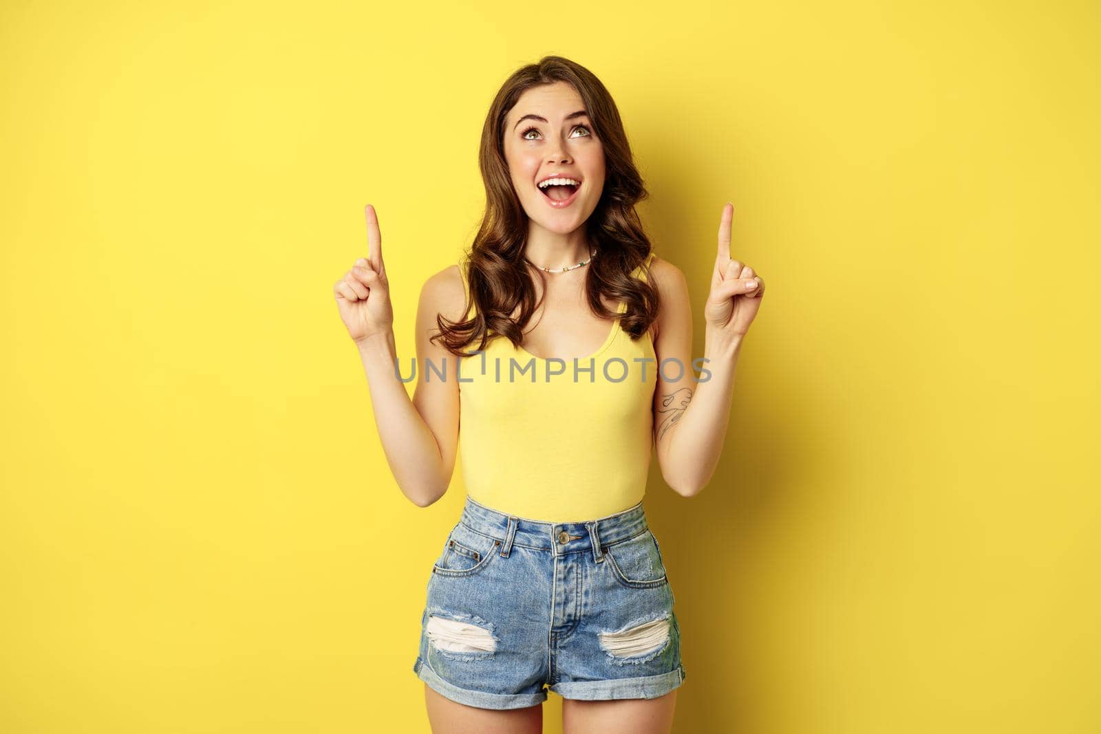Portrait of cheerful girl smiling, pointing fingers up, showing promo text, summer advertisement, standing against yellow background by Benzoix