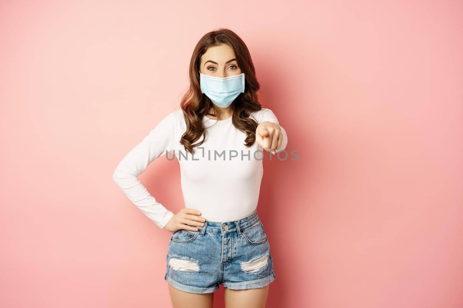 Its you. Happy smiling woman in medical face mask, pointing finger at camera, congratulating, praising, inviting people, seeing smth, standing over pink background by Benzoix