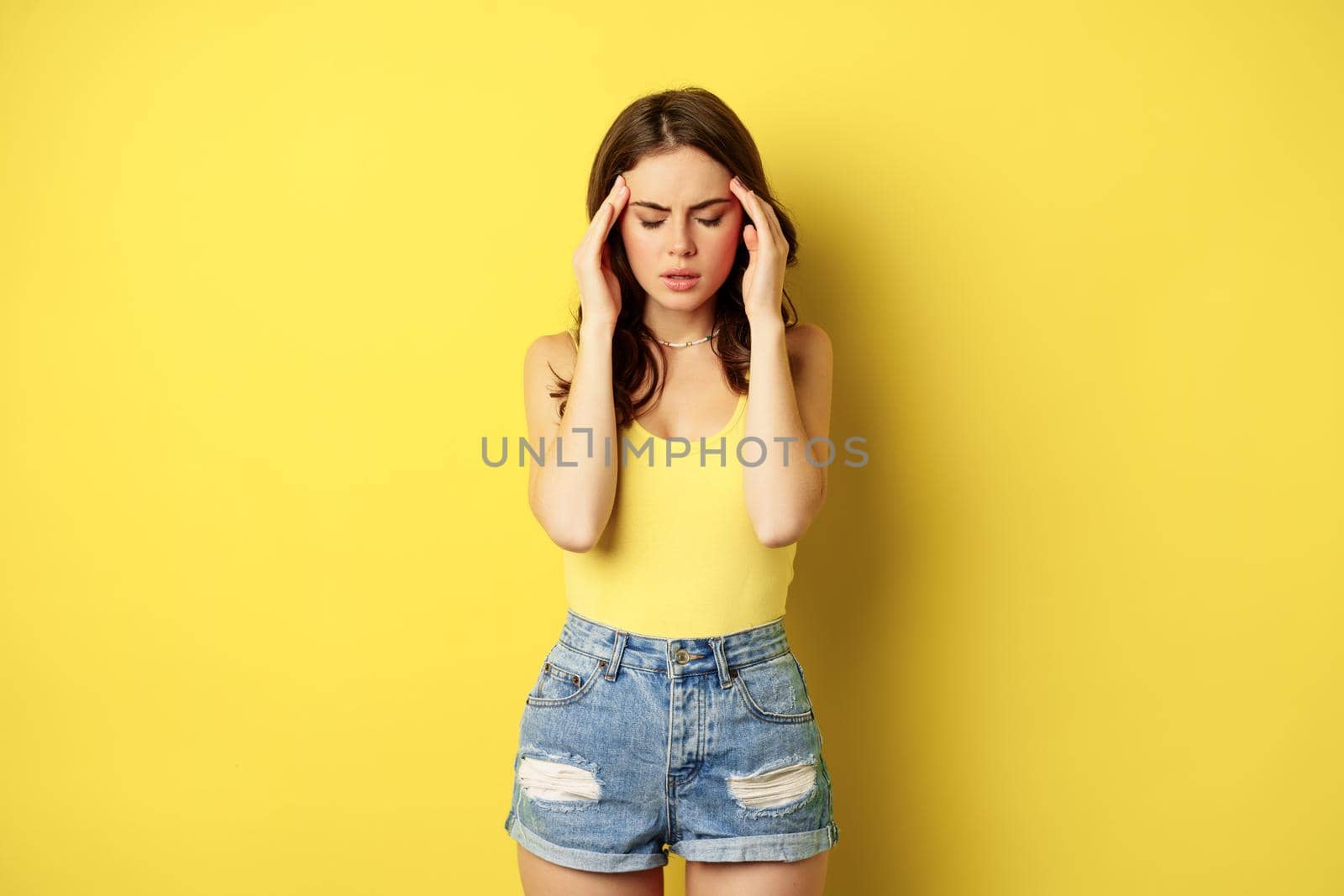 Stylish modern woman rubbing temples, having headache, migraine, frowning and grimacing from pain in head, standing against yellow background.