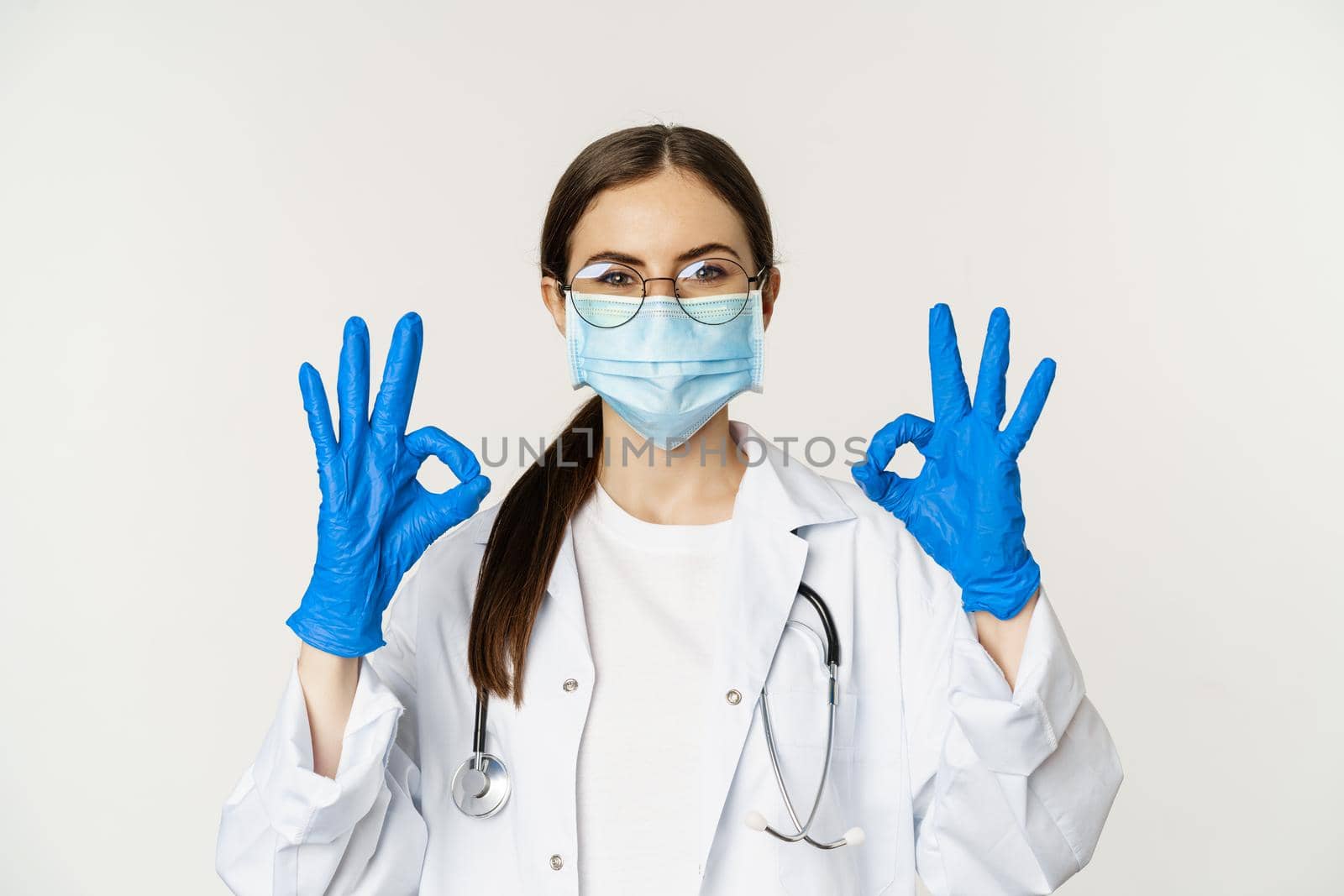Close up portrait of woman doctor, physician in face mask from coronavirus, showing okay sign in approval, recommending, praise and like smth good, standing over white background.