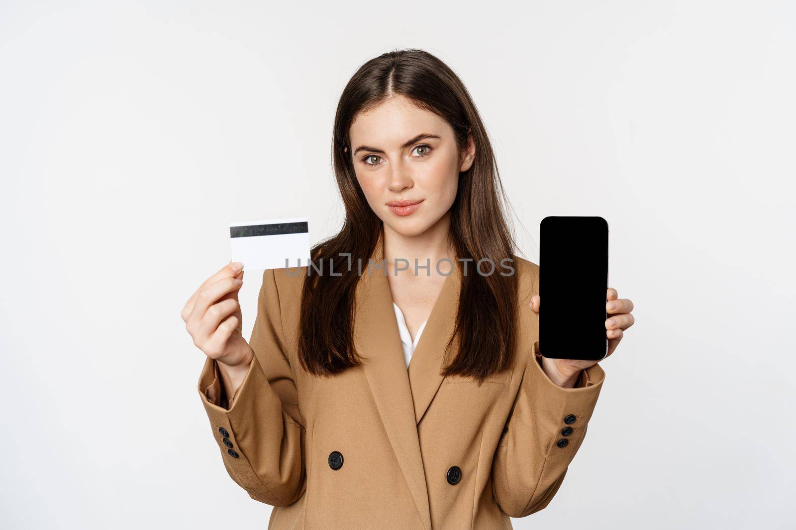Businesswoman showing mobile phone screen and credit card, demonstrating application on smartphone, standing in suit over white background.