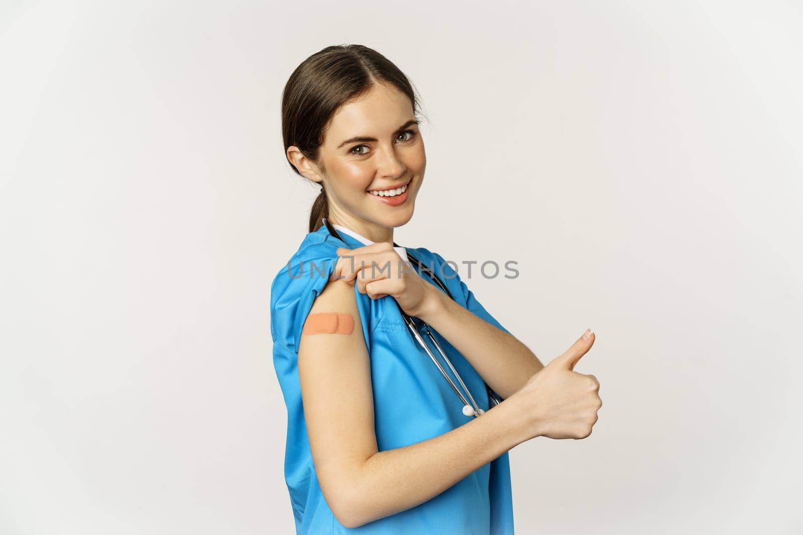 Smiling nurse, medical worker showing thumbs up and shoulder after vaccine, covid-19 vaccination campaign, standing over white background in scrubs.