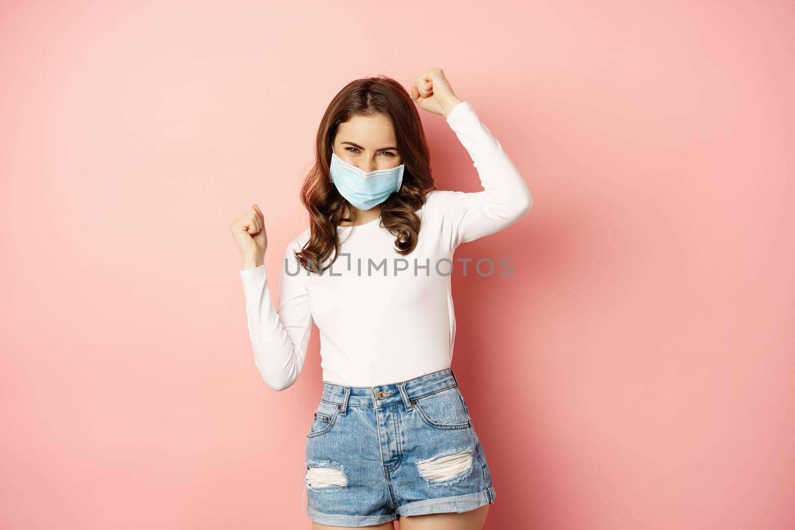 Enthusiastic brunette girl in medical face mask, dancing and laughing, celebrating victory, triumphing, winning smth, standing over pink background.