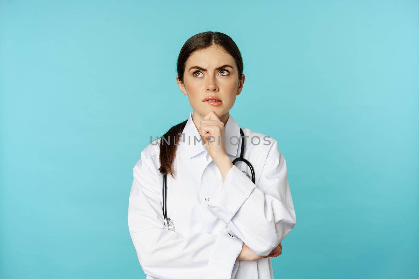 Young female doctor, hospital worker in white coat, thinking and looking away thoughtful, searching solution, standing over toquoise background.