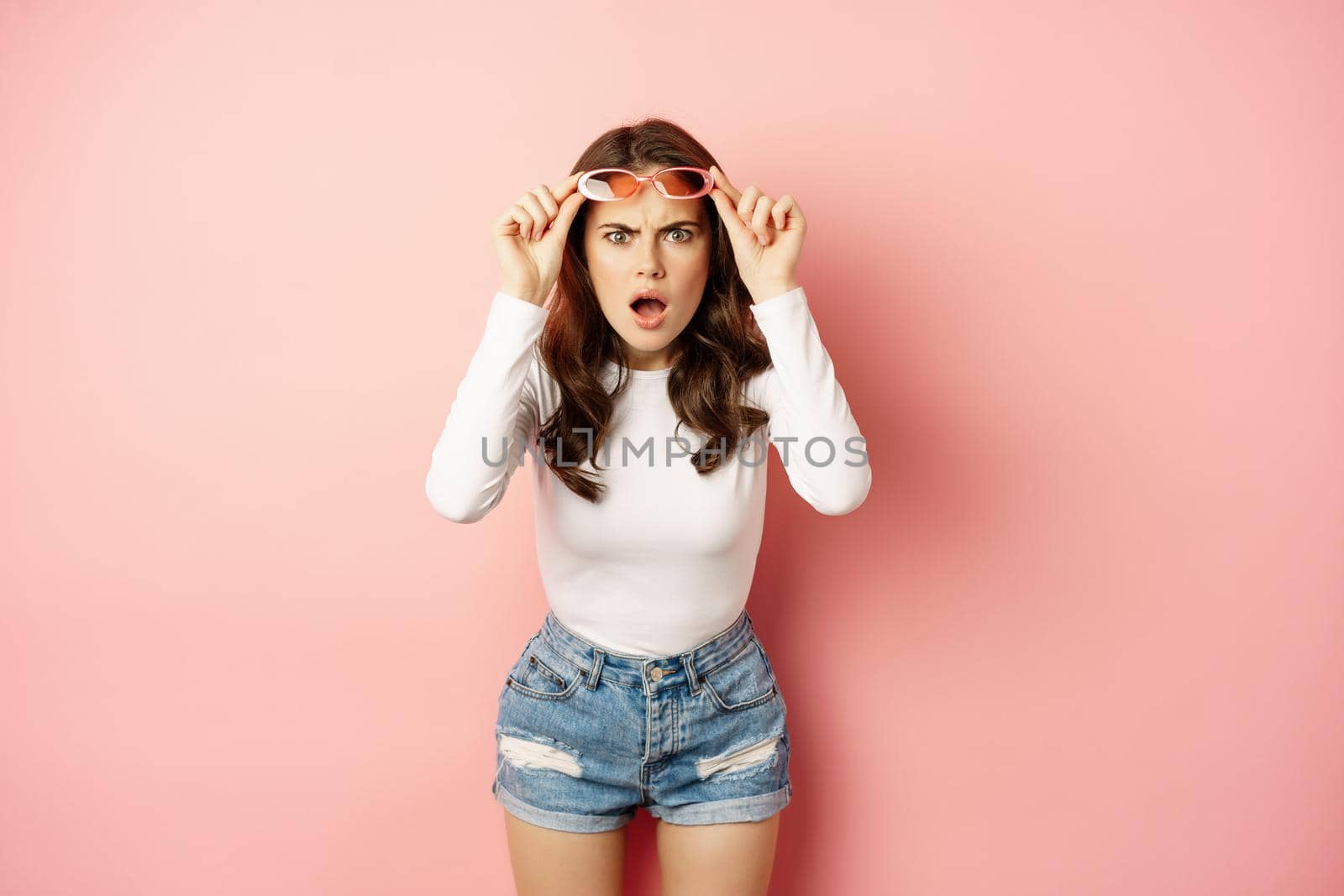 Portrait of shocked woman takes off sunglasses, looking confused, dont understand smth strange, reacting frustrated, standing over pink background by Benzoix