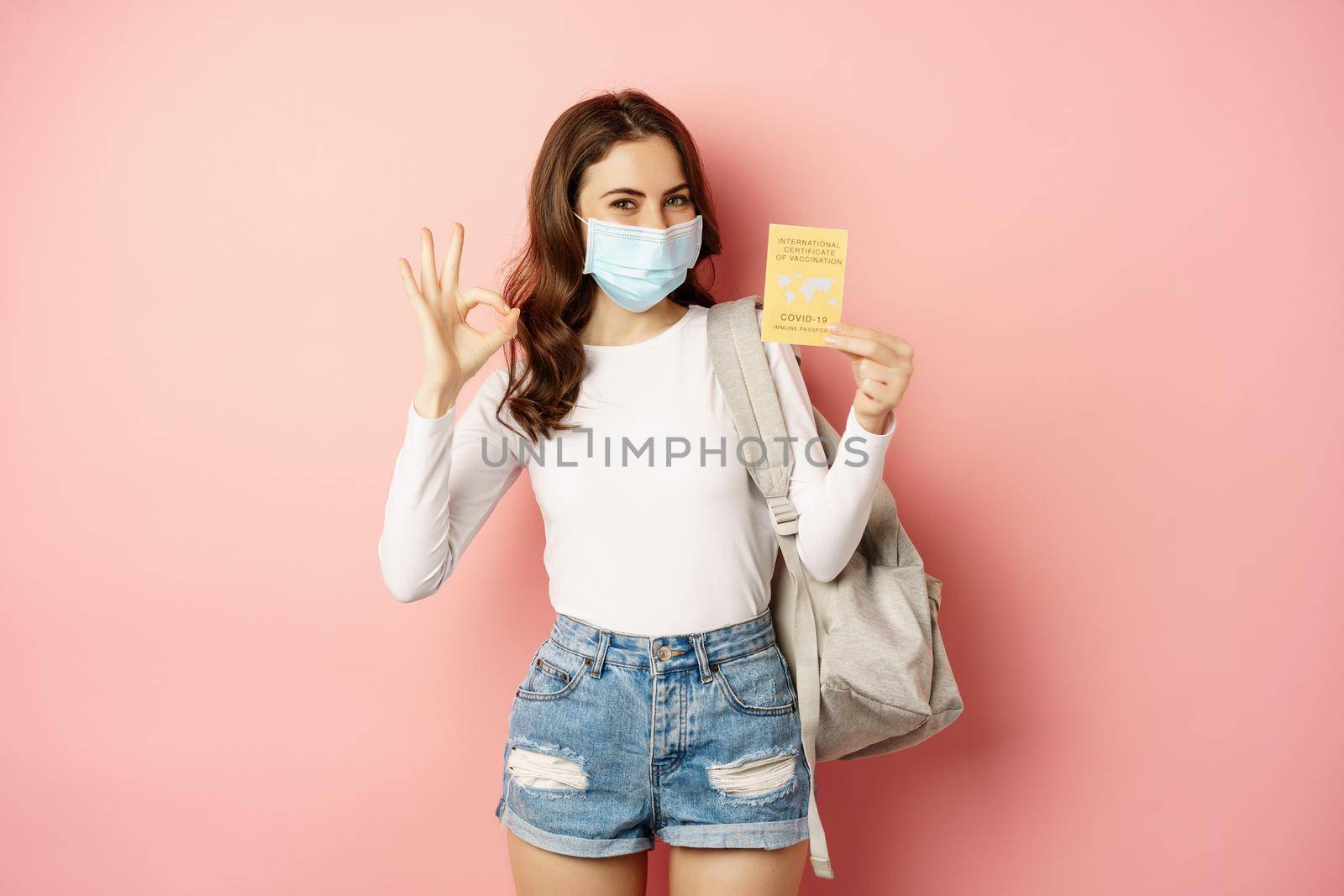 Young woman in medical mask, showing okay sign and covid vaccination certificate, travel during pandemic, going on holiday, standing over pink background.