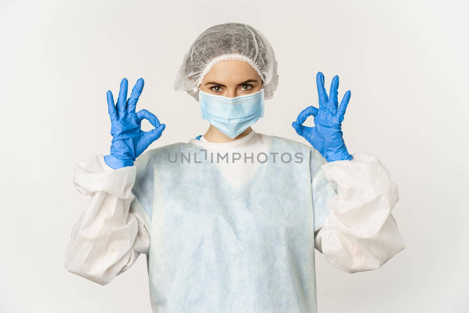 Image of healthcare worker, doctor in personal protective equipment from covid-19, showing okay ok sign, confident face in mask, standing over white background.