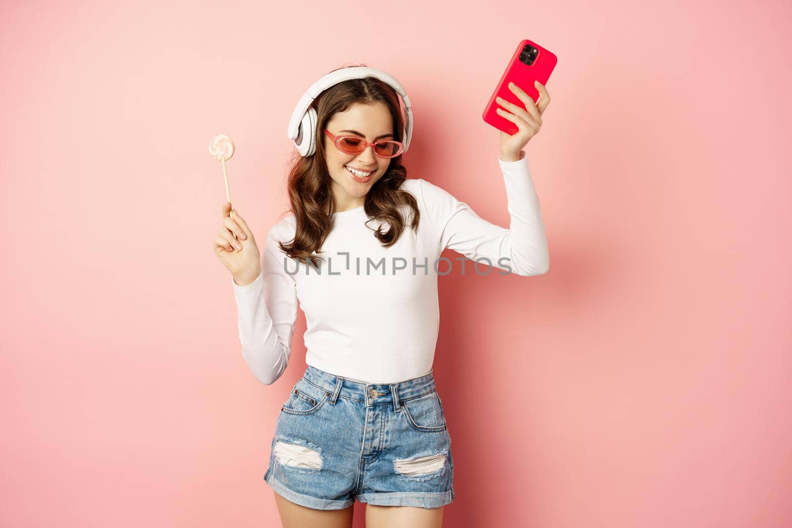 Dancing woman in sunglasses, listening music in headphones, holding lolipop and smartphone, laughing and smiling happy, standing over pink background by Benzoix