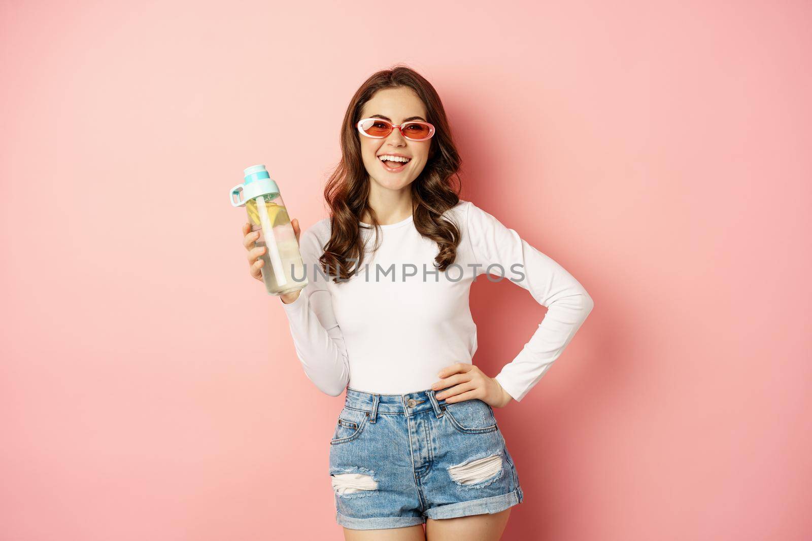 Stylish girl in spring outfit, wearing sunglasses, holding water bottle with lemon, healthy drink, laughing and smiling, standing over pink background by Benzoix