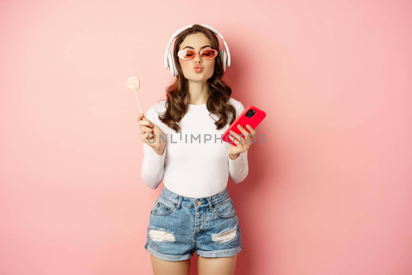 Stylish brunette woman dancing in headphones with mobile phone, listening music in earphones, licking lolipop, standing over pink background.