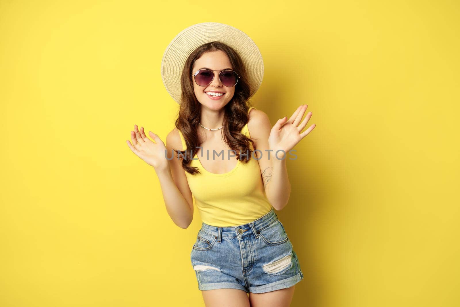 Happy sexy young woman on vacation, wearing straw hat, sunglasses, laughing and smiling, standing over yellow background.