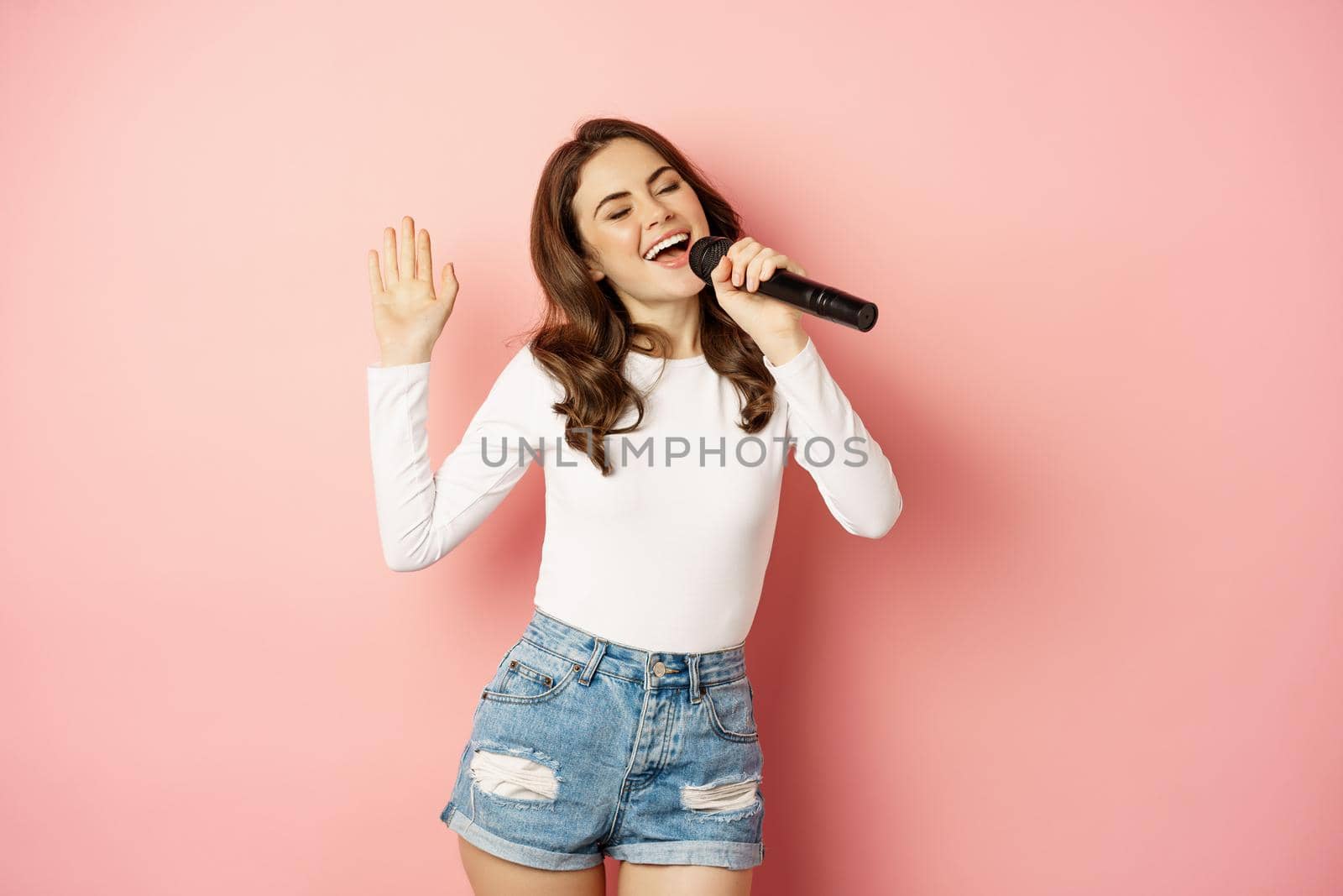 Party girl. Happy young woman singing in microphone, performing song, having fun at event, standing over pink background.