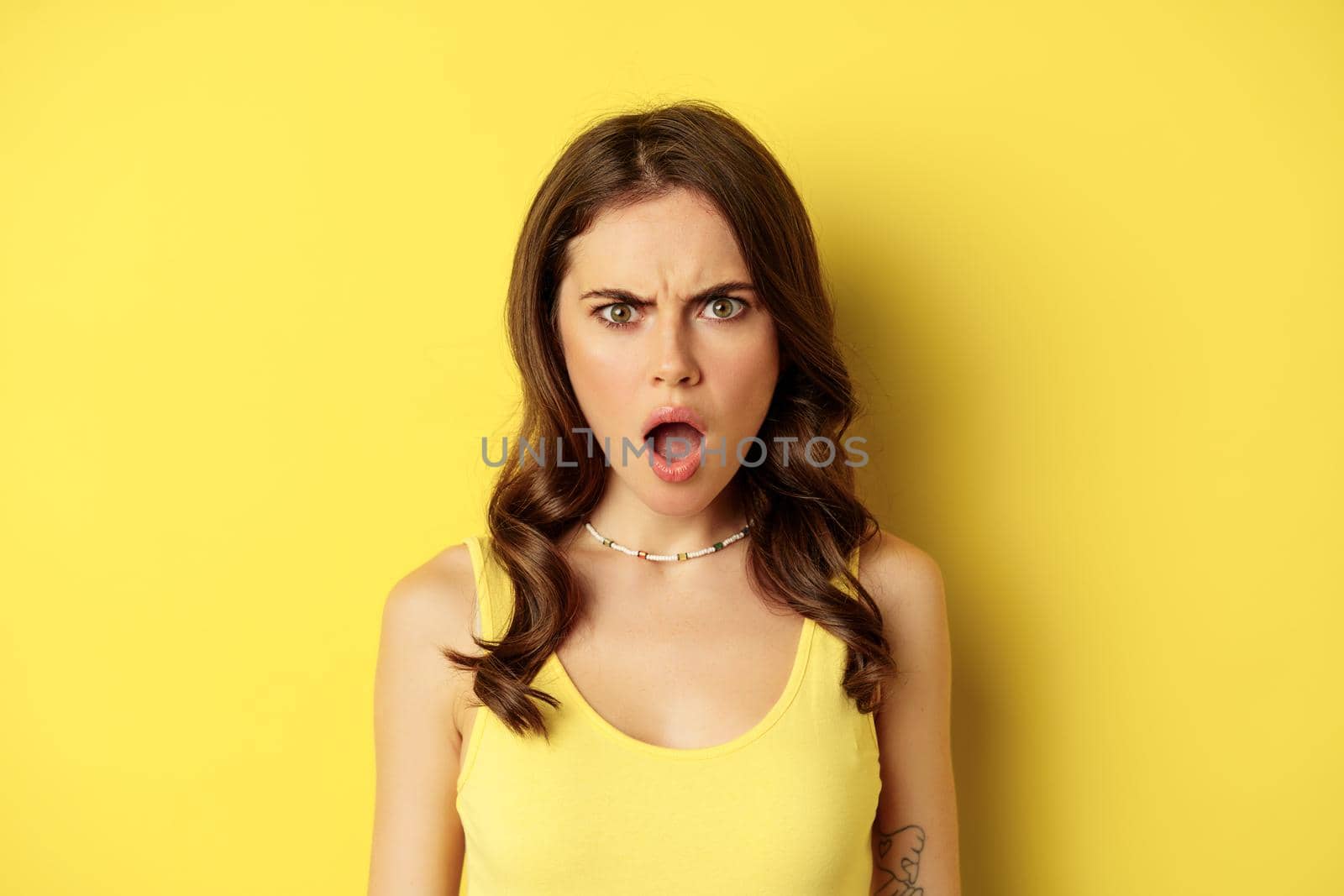 Close up portrait of shocked young woman drop jaw, gasping and looking frustrated, standing against yellow background. Copy space