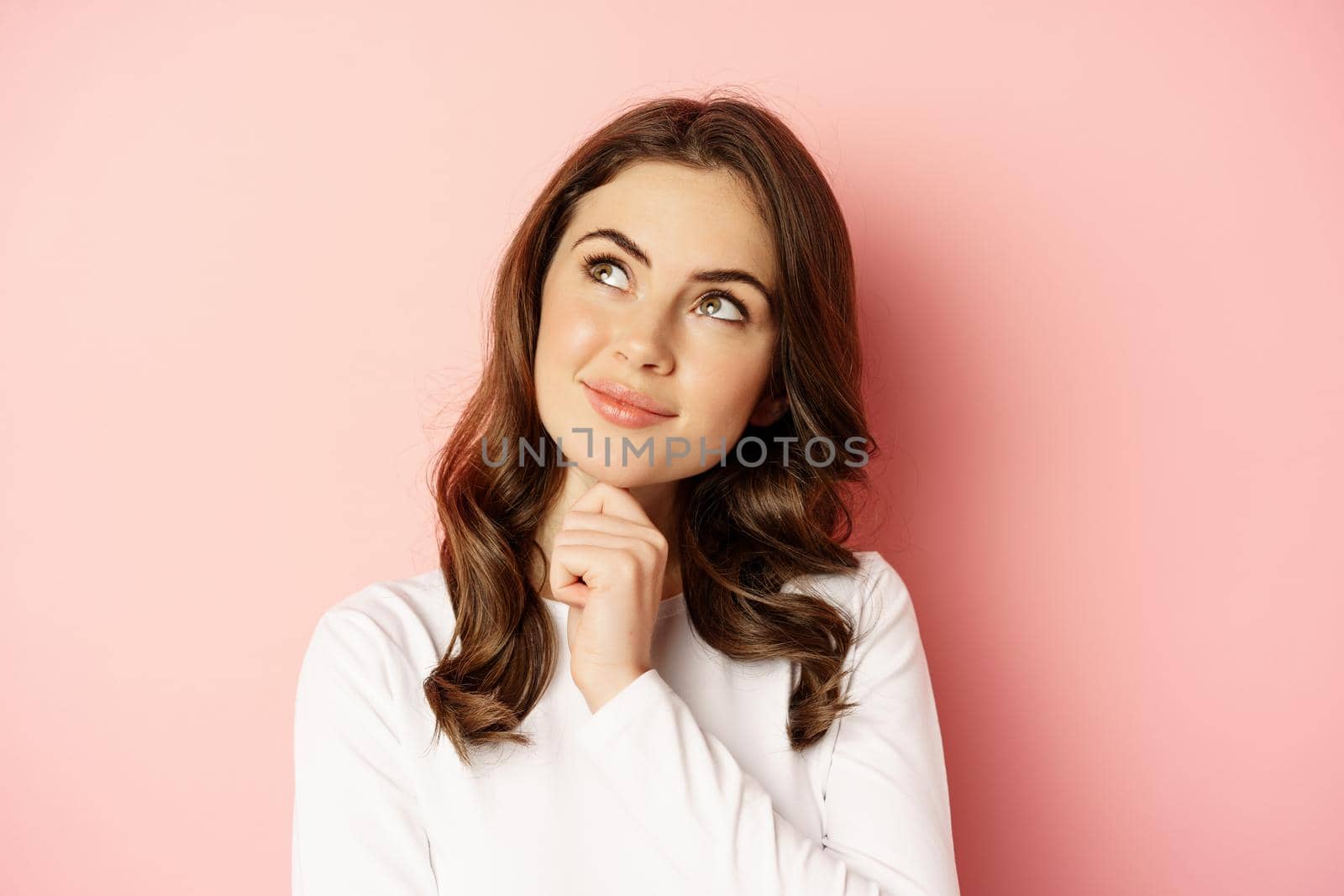 Close up portrait of coquettish smiling woman, glamour girl thinking, looking thoughtful, standing over pink background.