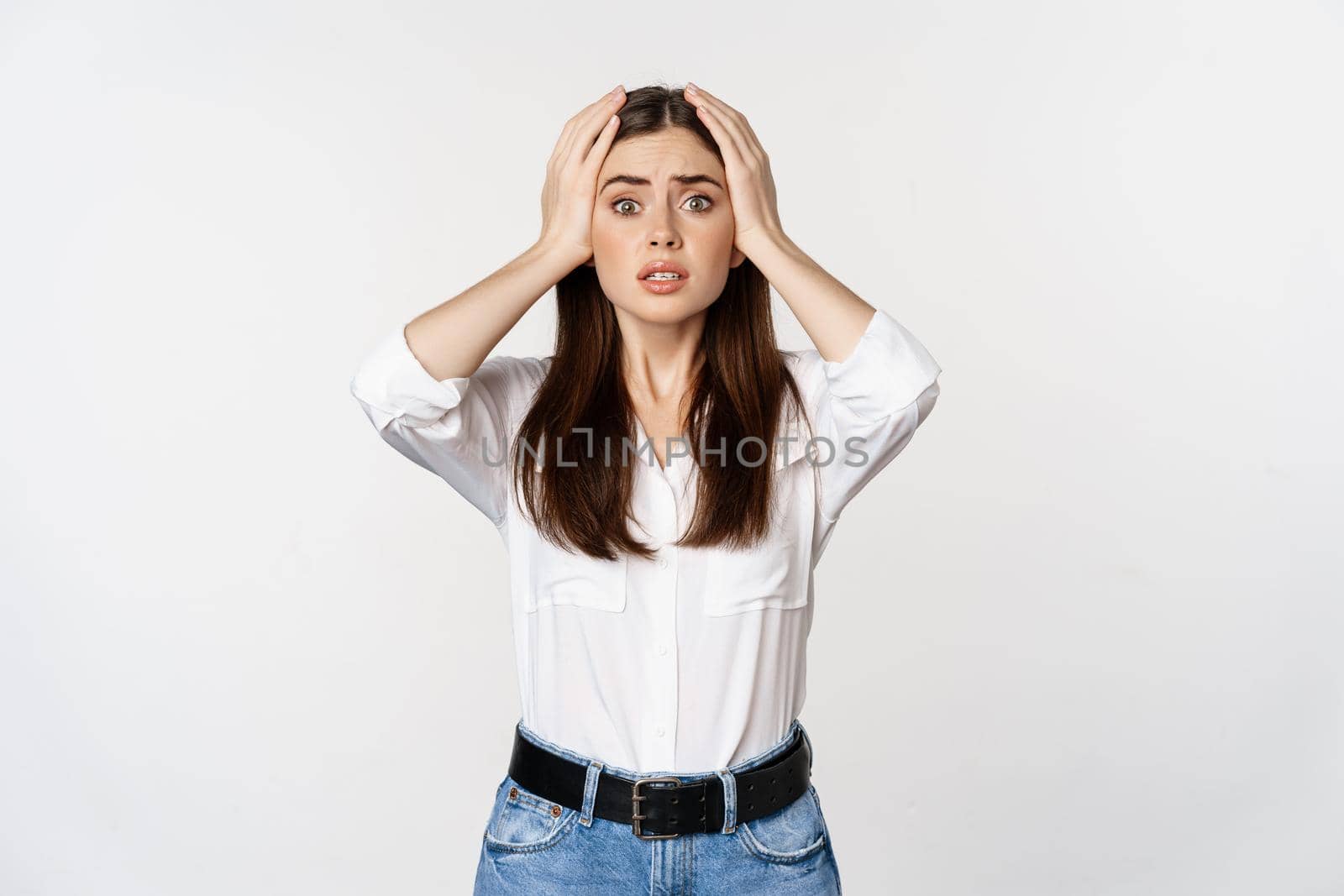 Worried and shocked young woman, looking stunned at disaster, holding hands on head, panicking, standing over white background.