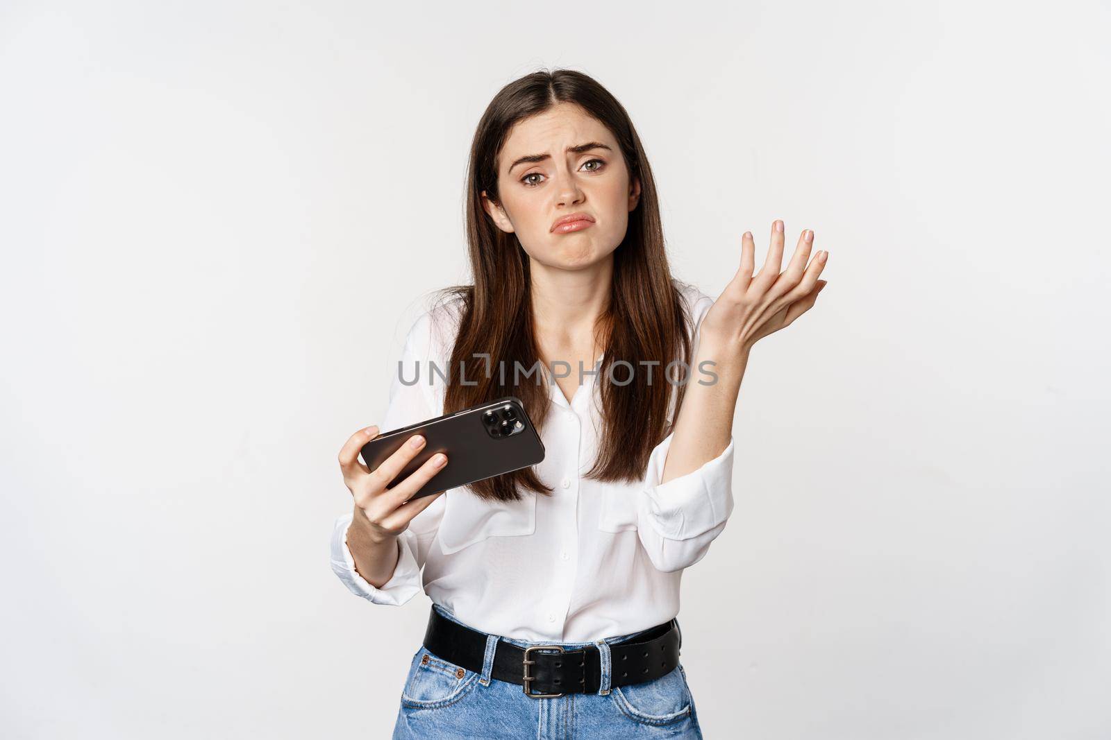 Sad woman losing in mobile video game, looking upset and disappointed at smartphone, sulking, standing over white background.