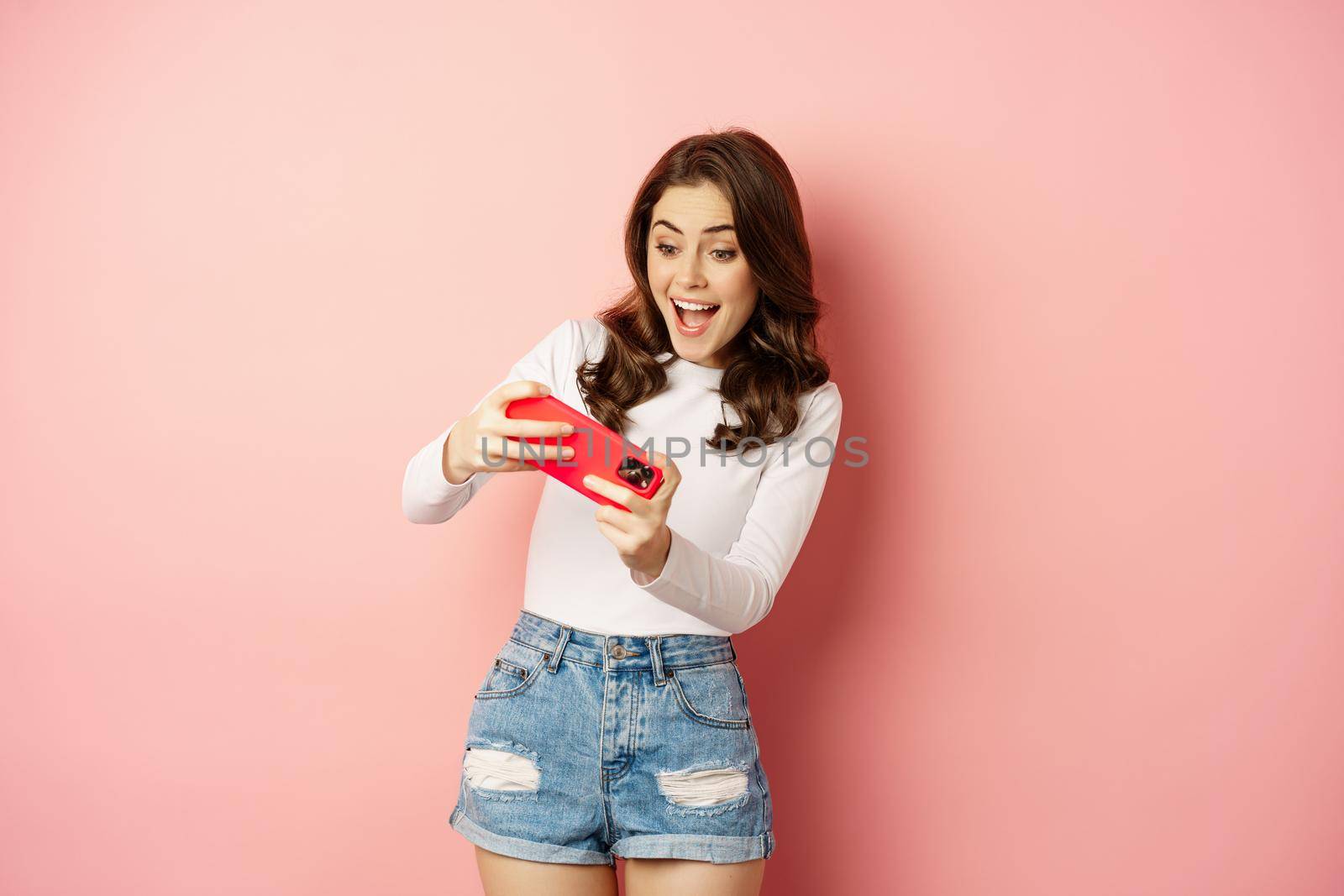Happy beautiful girl playing mobile video game, holding smartphone horizontally, watching on cellphone with excited face, pink background.