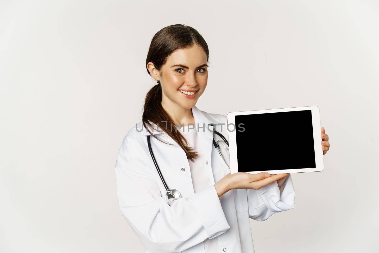 Image of woman doctor, female healthcare worker showing online medical website, digital tablet screen and smiling, standing in white coat over white background.