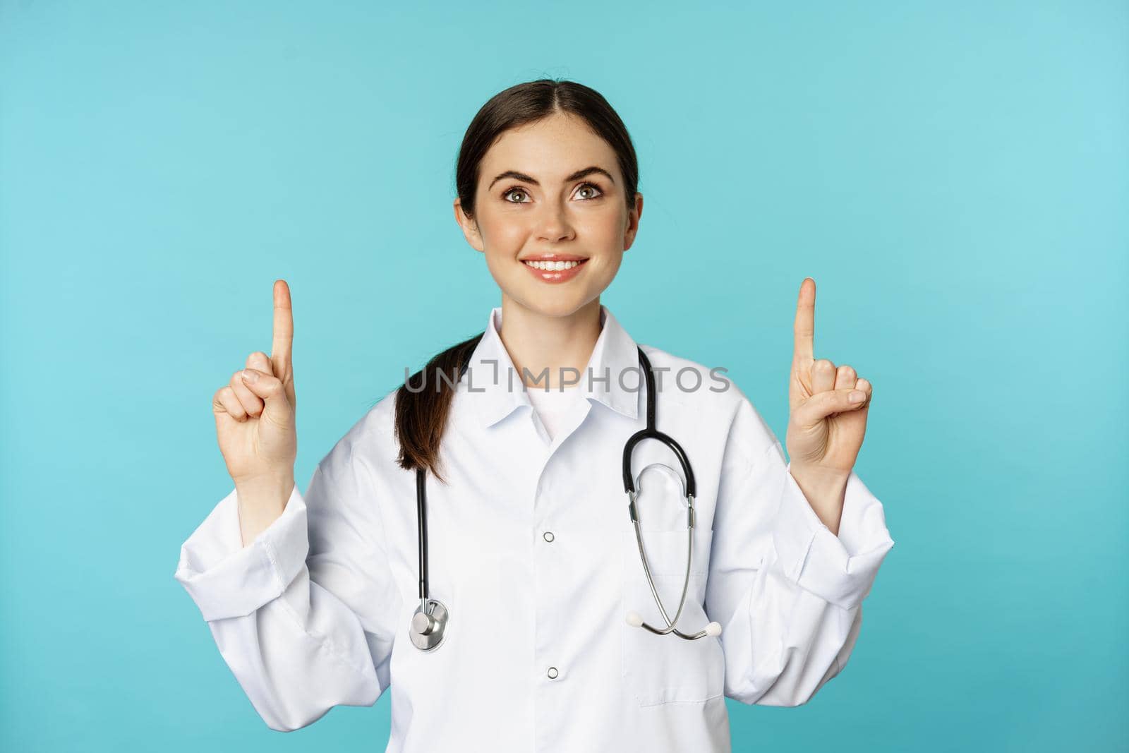 Enthusiastic medical worker, young woman doctor in white coat, stethoscope, showing advertisement, pointing fingers up, standing over torquoise background.
