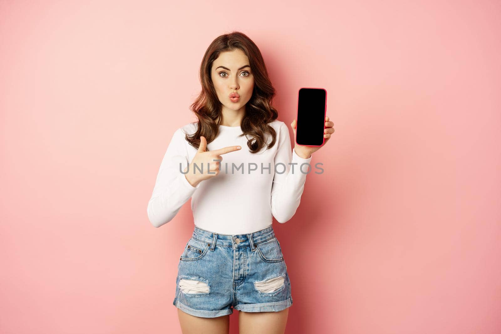 Surprised brunette girl pointing at mobile phone app screen, showing smartphone advertisement, online shopping offer, standing amazed against pink background.