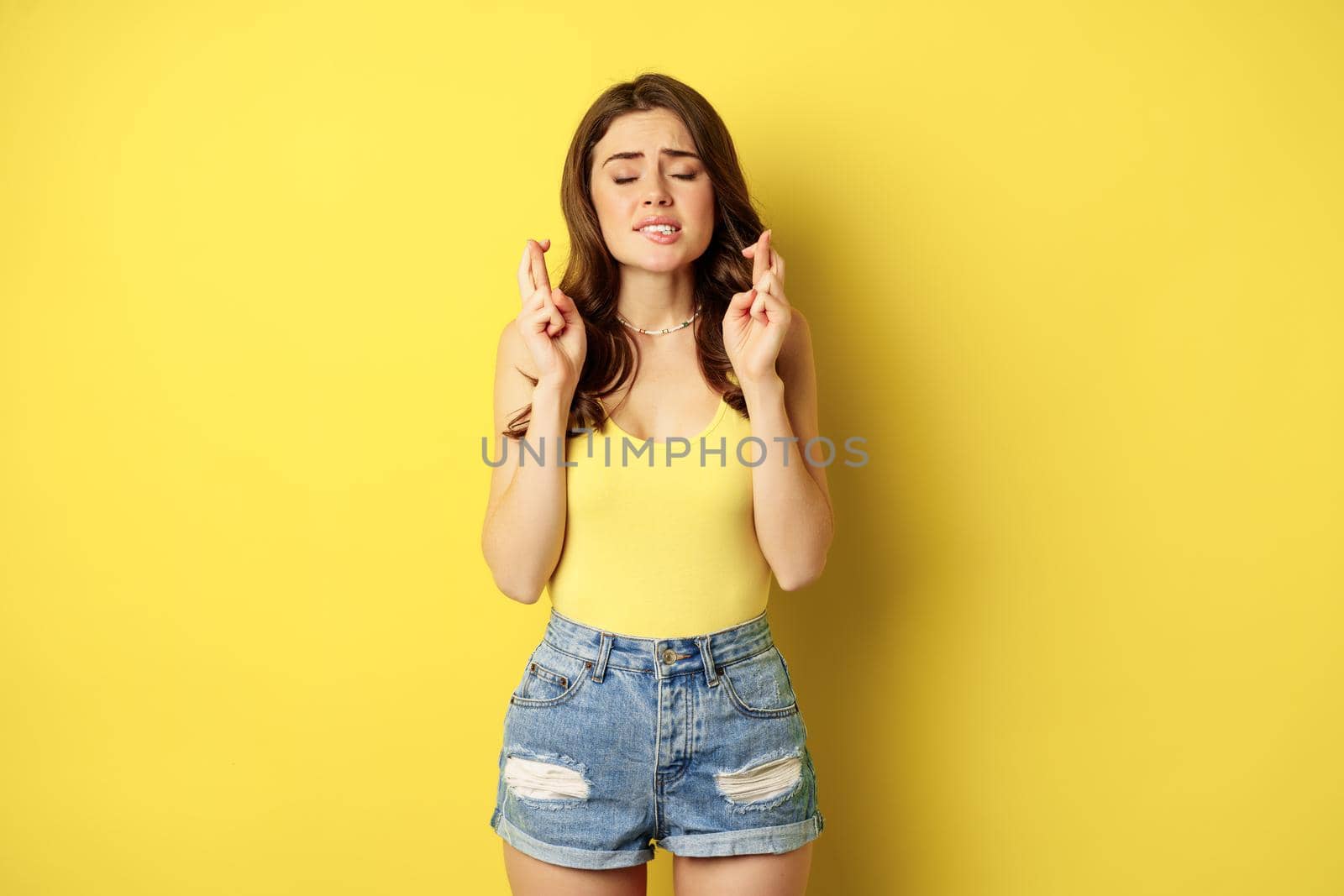 Hopeful young beautiful woman praying, believe, hoping to receive smth, cross fingers for good luck, anticipating, standing over yellow background.