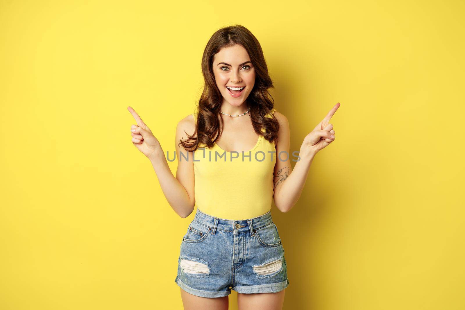 Portrait of stylish young female model, showing advertisement, pointing at logo or banner, demonstrating two ways, choices in store, standing over yellow background by Benzoix