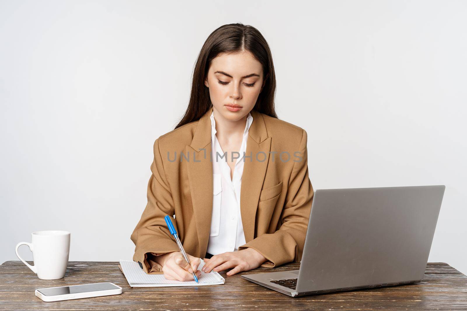 Image of businesswoman working in office, sitting at table with laptop, writing documents, posing in formal suit over white background.