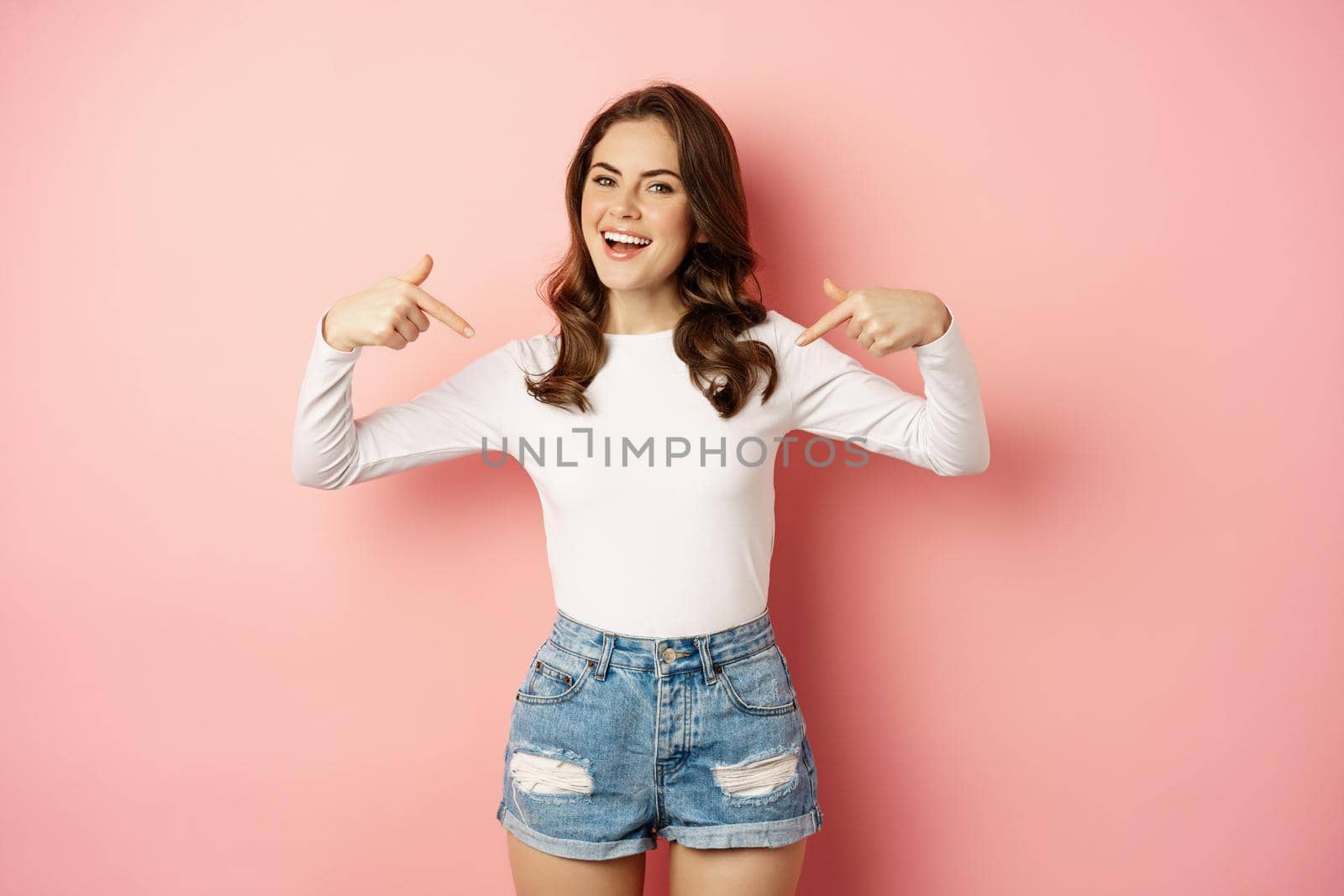 Self-confident young happy woman, stylish girl pointing at herself, smiling satisfied, self-promoting, talking about her, standing against pink background.