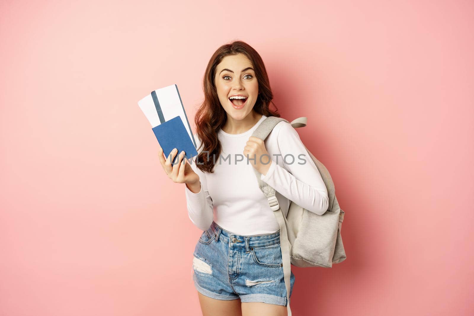 Vacation and holiday. Beautiful girl going on trip, holding passport with airplane tickets, holding backpack, travelling, standing over pink background by Benzoix