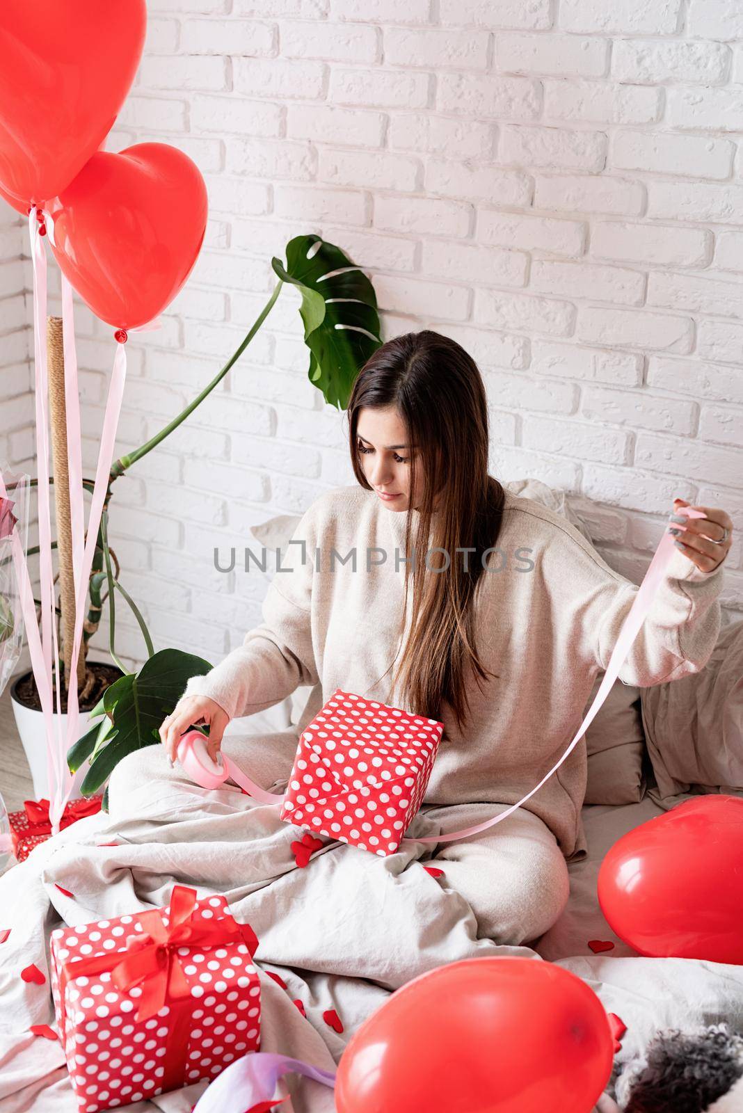 Valentine's day, Women's day. Young caucasian brunette woman sitting in the bed celebrating valentine day wrapping the gifts