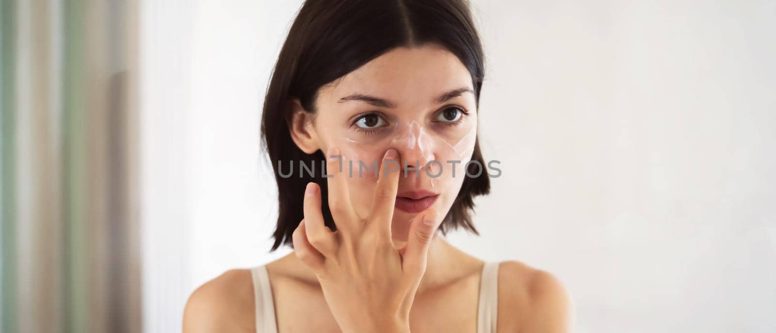 A young girl applies a moisturizing cream mask near the mirror in the bathroom and holds a jar of balm in her hand. A woman takes care of the health of her skin, uses cosmetic masks.