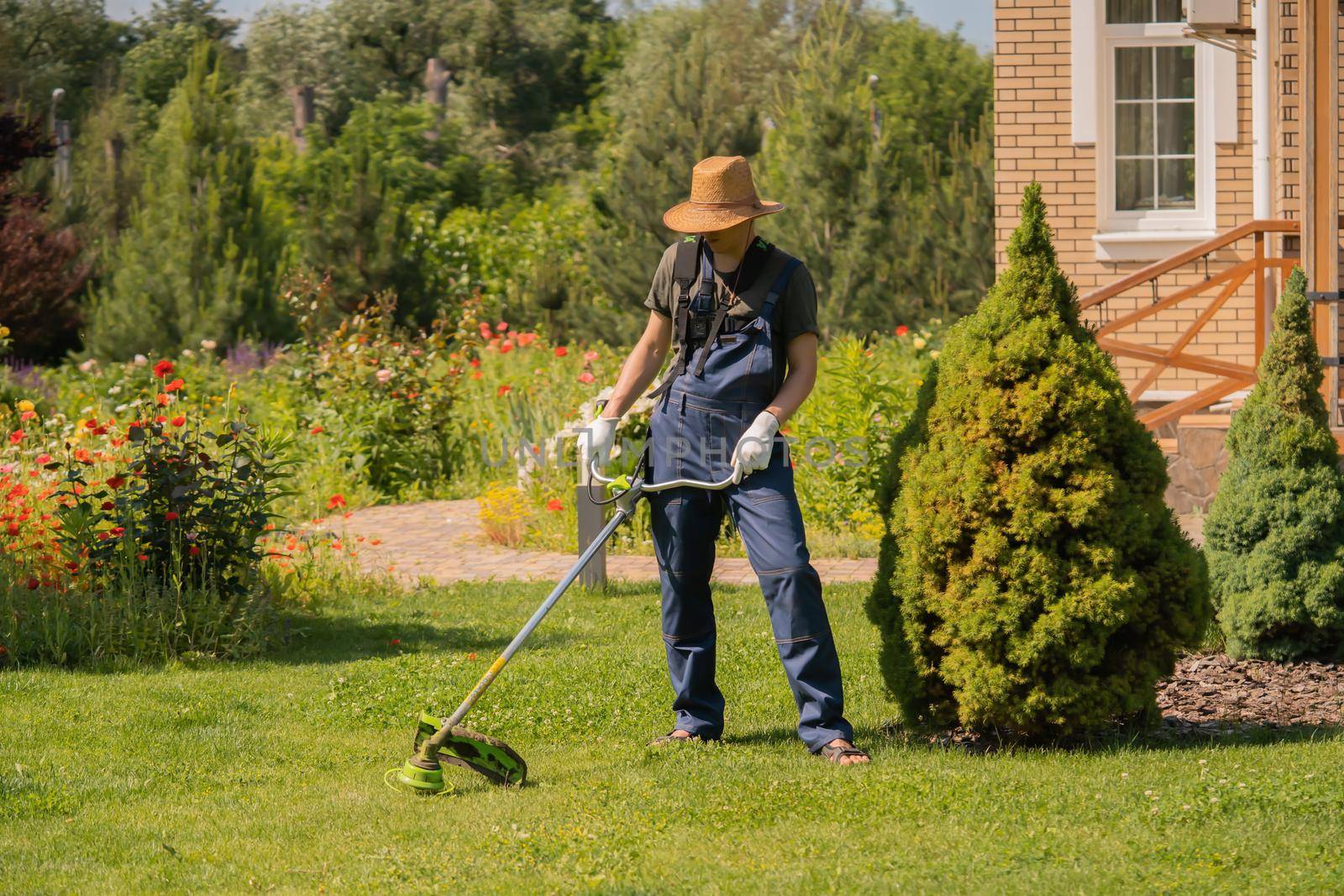 A young man in a straw hat is mowing a lawn with a lawn mower in his beautiful green floral summer garden. A professional gardener with a lawnmower cares for the grass in the backyard.