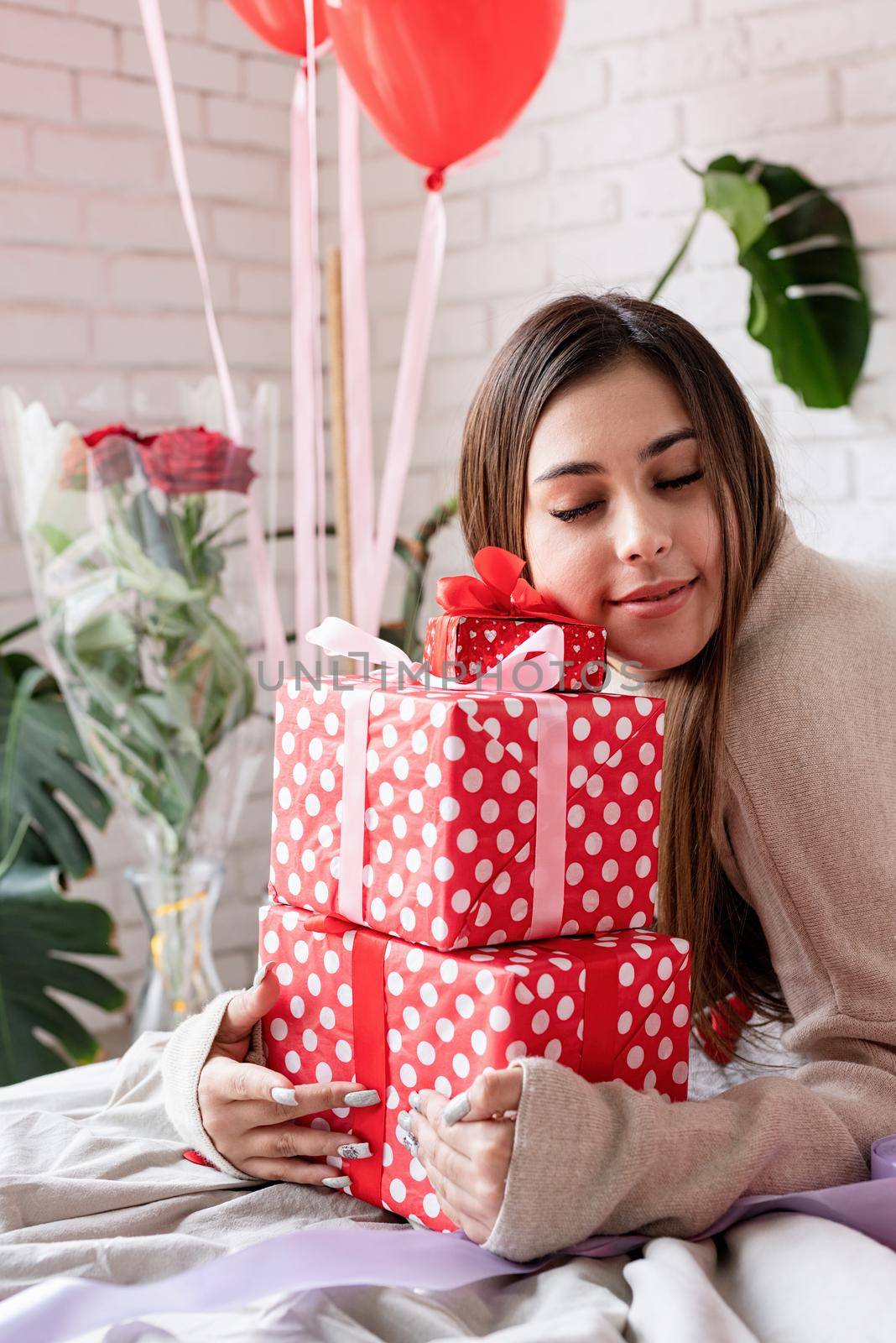 Young woman sitting in the bed celebrating valentine day embracing pile of gifts by Desperada