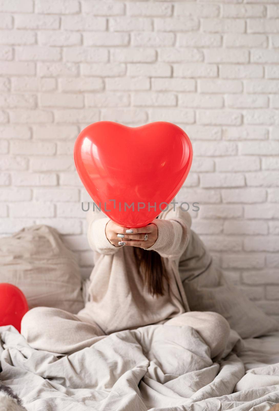 Young brunette woman sitting in the bed celebrating valentine day holding red heart balloons by Desperada