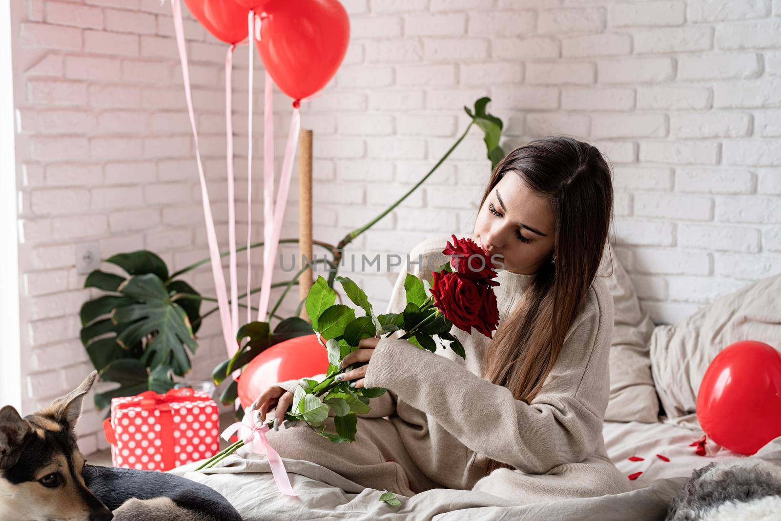 Valentine's day, Women's day. Young caucasian brunette woman sitting in the bed celebrating valentine day smelling red roses