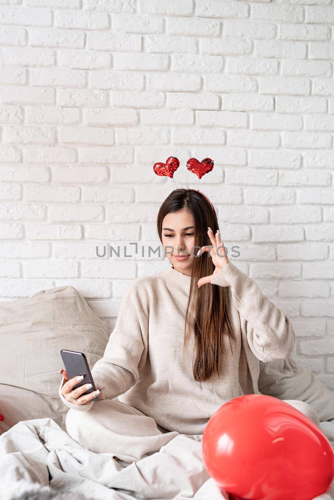 Young funny woman sitting in the bed celebrating valentine day chatting using mobile phone by Desperada
