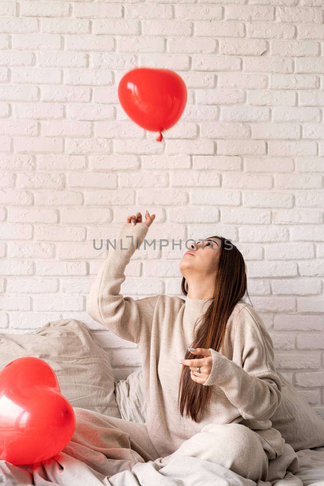 Valentine's day, Women's day. Young brunette woman sitting in the bed celebrating valentine day holding red heart balloons