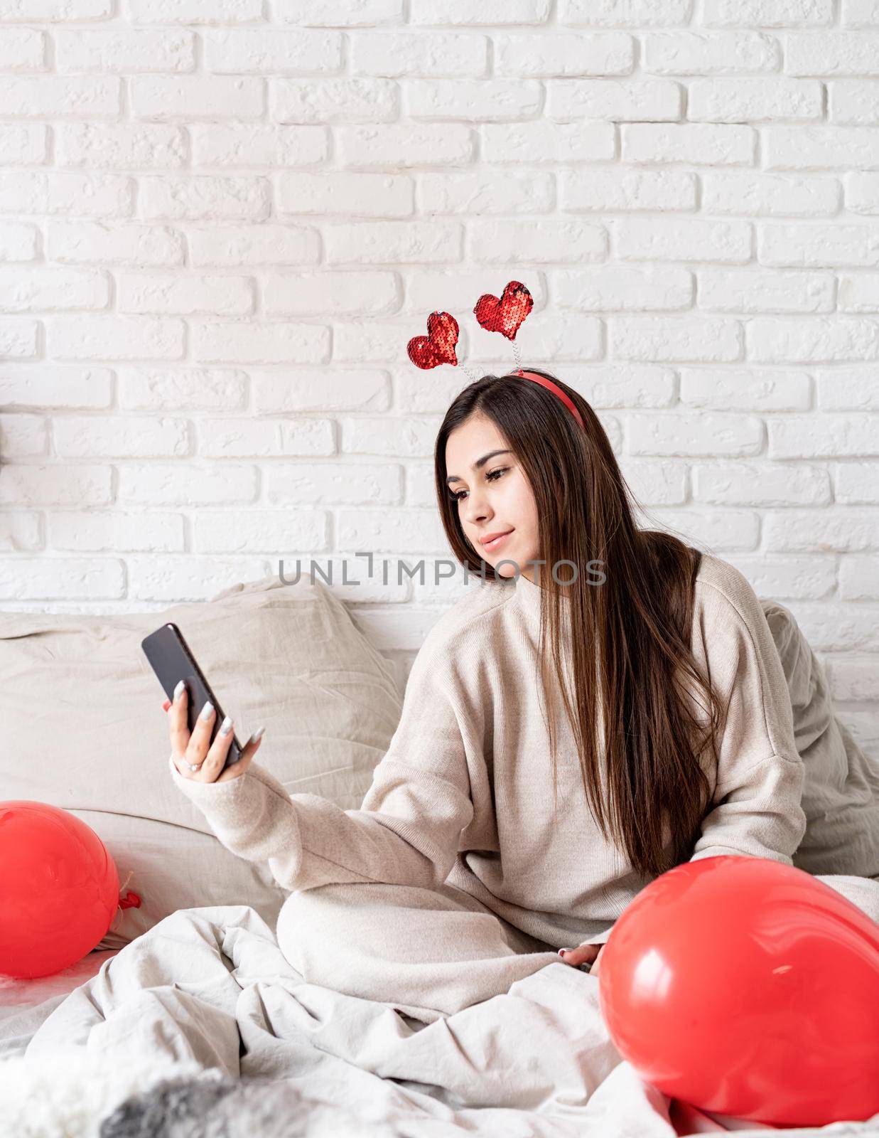 Valentine's day, Women's day. Young funny woman sitting in the bed celebrating valentine day chatting using mobile phone