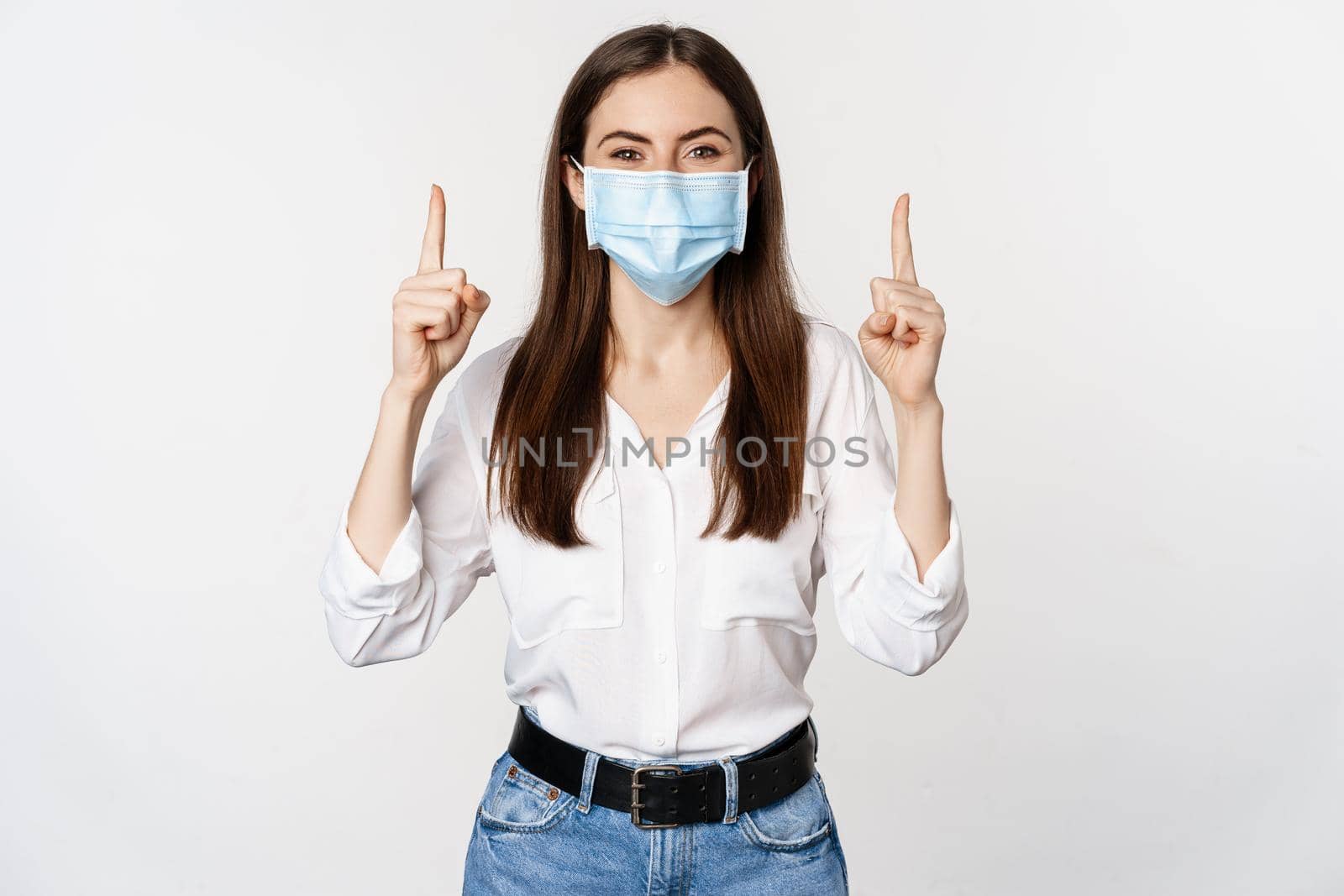 Portrait of corporate woman in face medical mask from coronavirus, pointing fingers up, showing advertisement, white background.