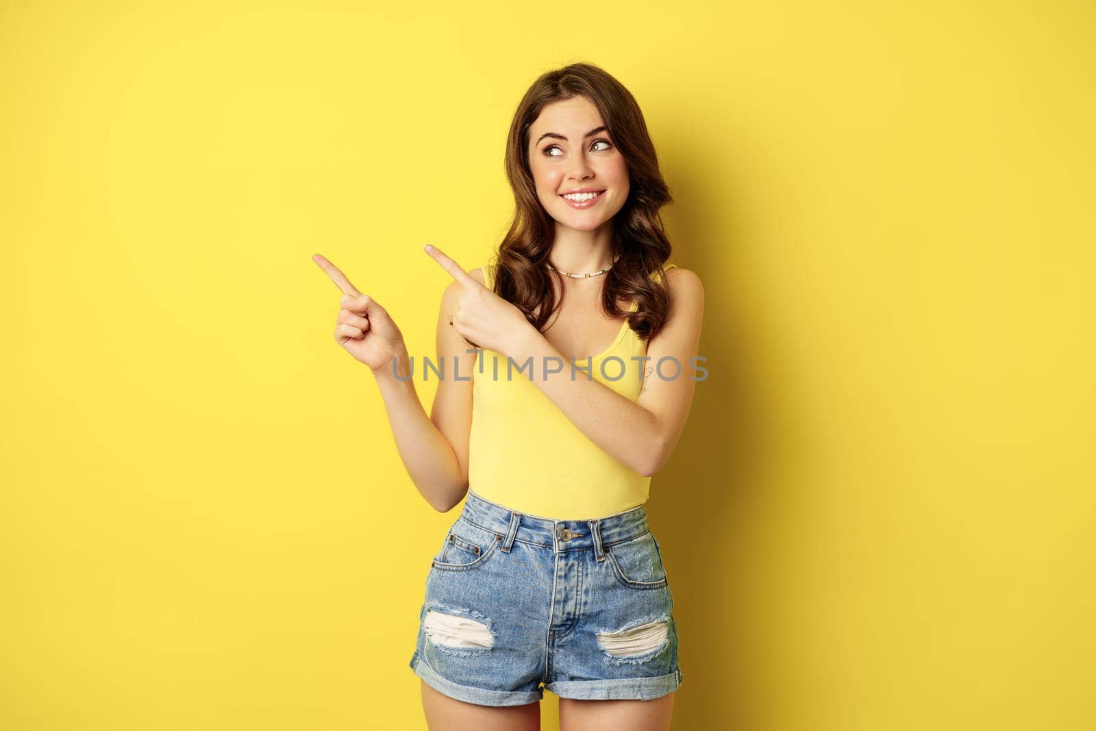 Portrait of stylish summer girl, brunette woman showing way, pointing fingers left, advertising, standing over yellow background. Copy space